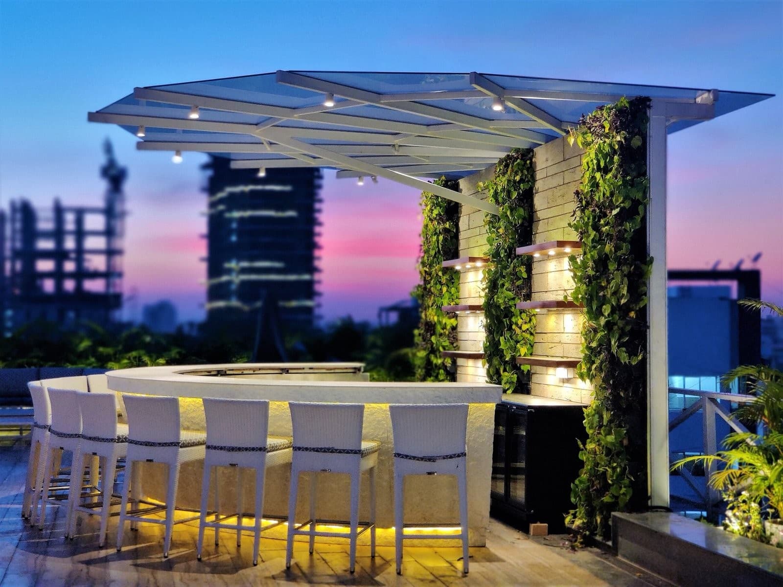 A rooftop bar at dusk: a white pergola over a lit counter, planted walls and the city skyline beyond.